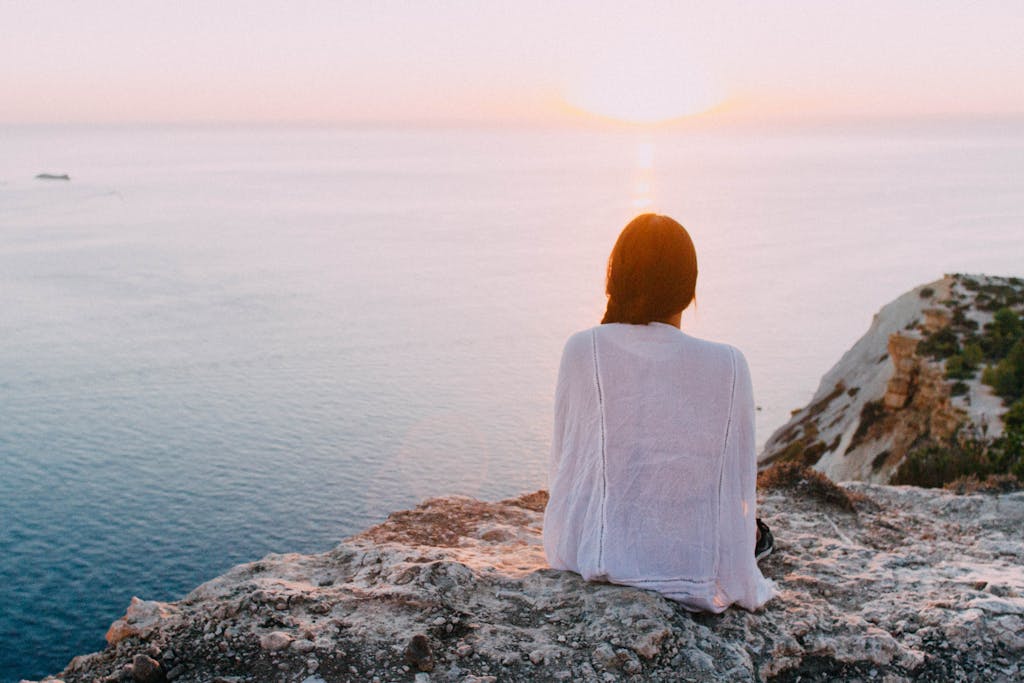 a serene and peaceful woman sitting on a rocky cliff staring at the ocean during sunset self development personal growth calm 